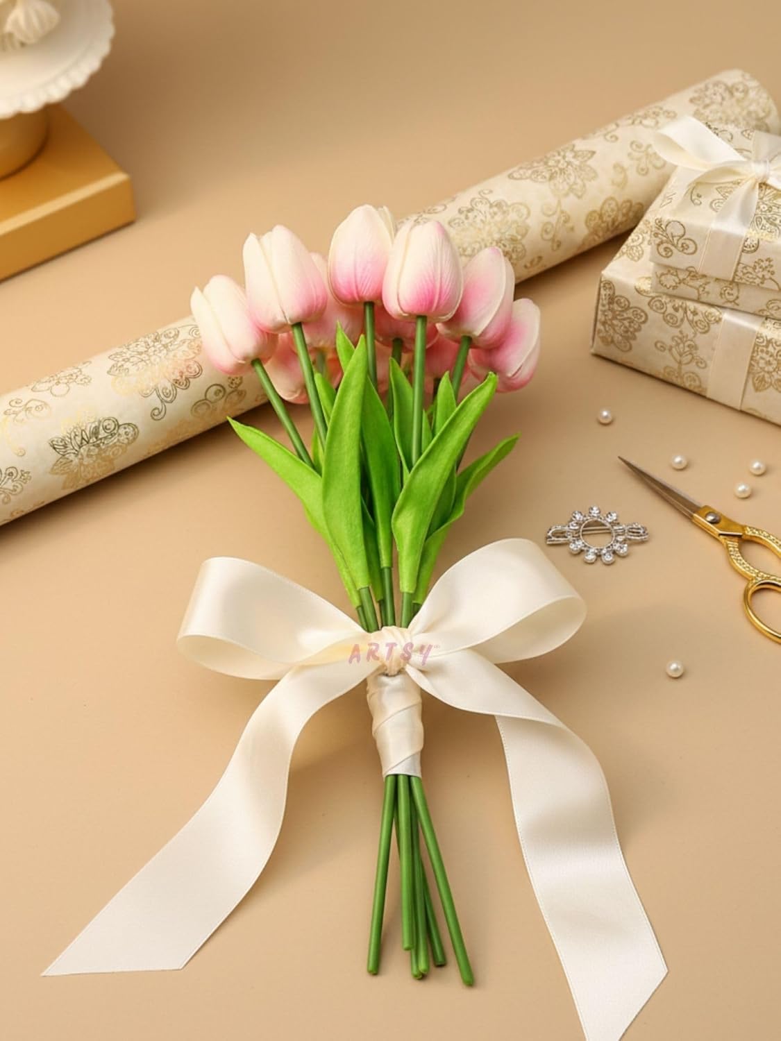 Bouquet of baby pink tulips with a white ribbon on a beige surface with decorative items.