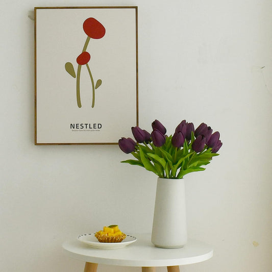 Vase of purple tulips on a table with a framed artwork in the background.